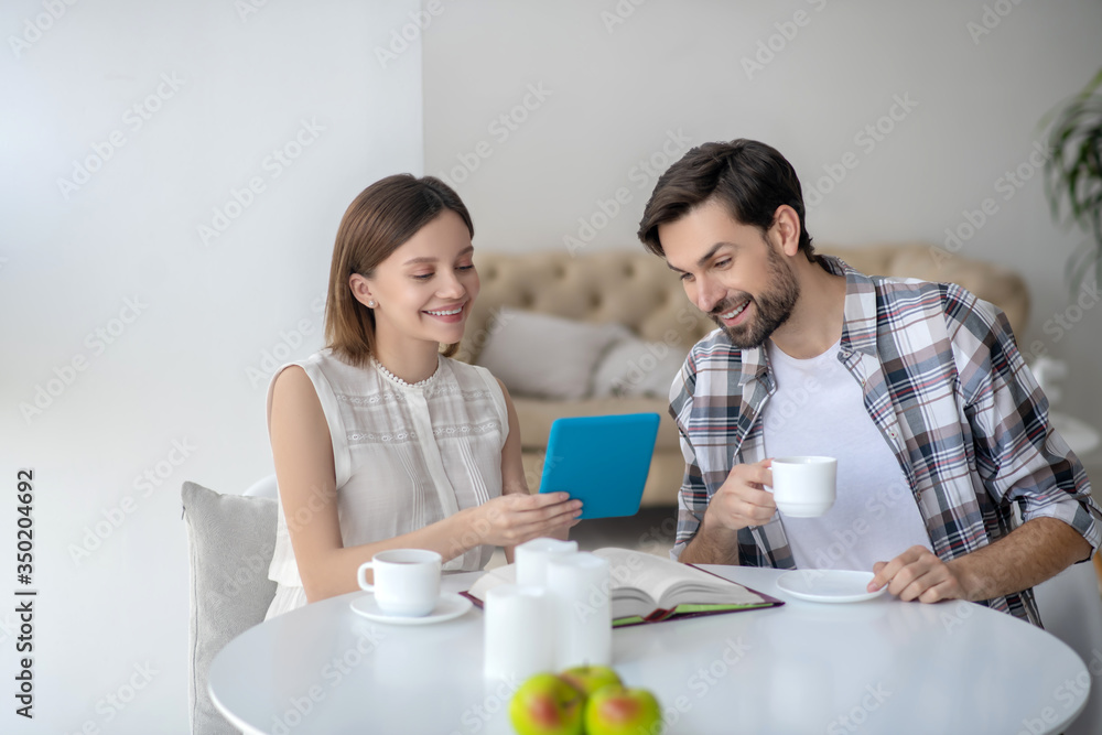 Cute young woman in a white dress holding a tablet in her hand and showing something to her husband