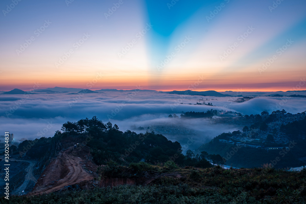 Fototapeta premium Mountains in fog at beautiful morning in autumn. Landscape with Langbiang mountain valley, low clouds, forest, colorful sky , city illumination at dusk.