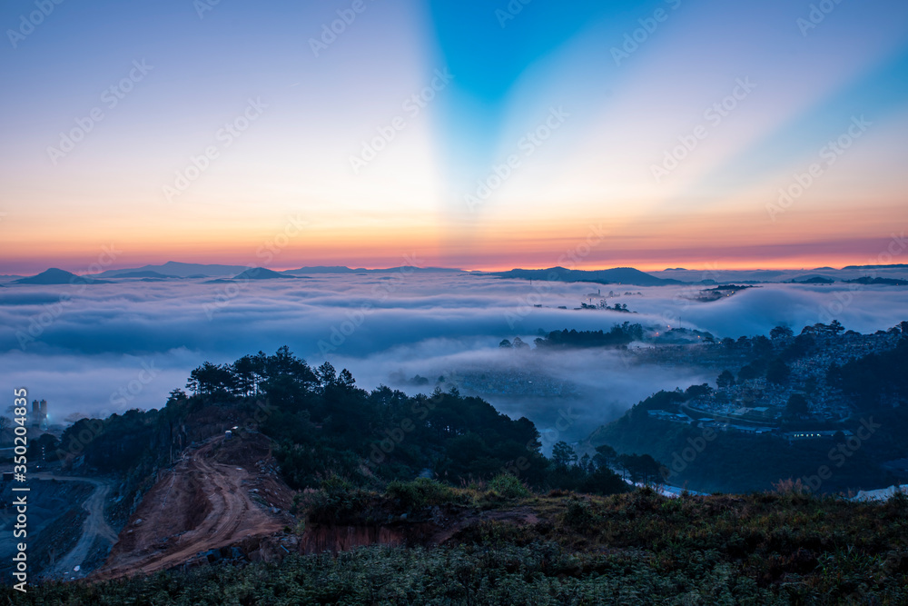 Mountains in fog at beautiful morning in autumn. Landscape with Langbiang mountain valley, low clouds, forest, colorful sky , city illumination at dusk.