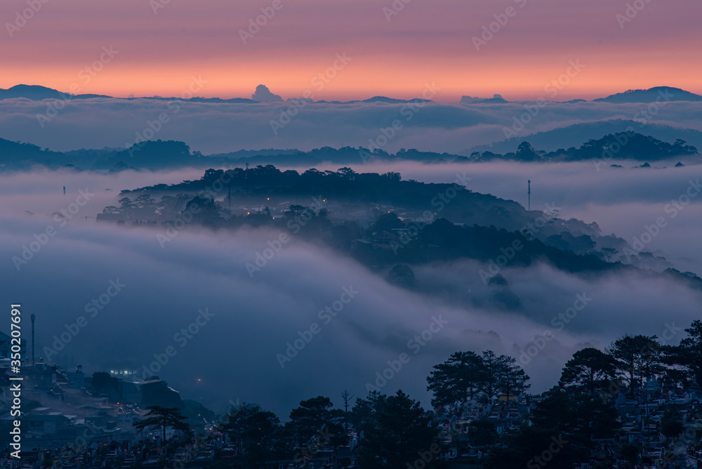 Mountains in fog at beautiful morning in autumn. Landscape with Langbiang mountain valley, low clouds, forest, colorful sky , city illumination at dusk.