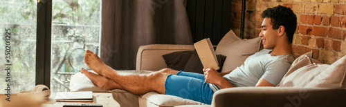 smiling mixed race man reading book on sofa during quarantine, website header