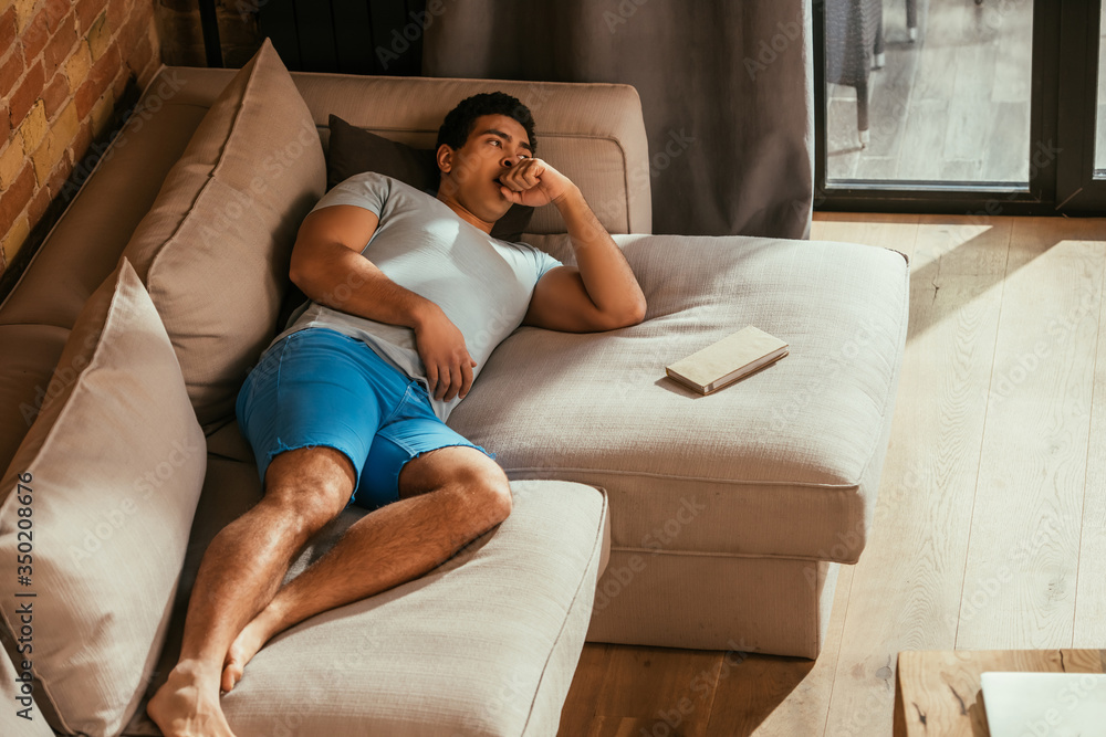 Obraz premium handsome mixed race man lying on sofa with book at home during quarantine
