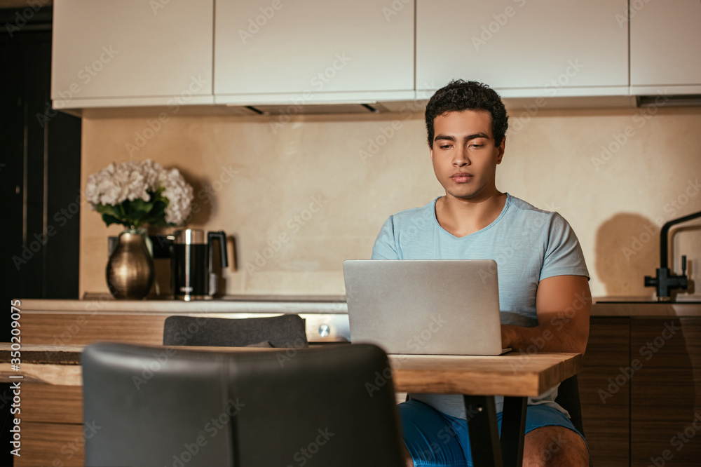 handsome male mixed race freelancer working with laptop on kitchen during quarantine