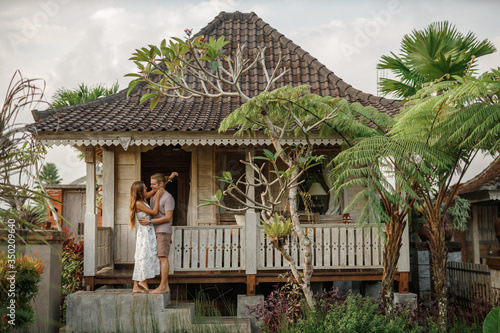 Young lovely couple wearing natural clothes standing on the porch of wooden house, hugs and smiles. Honeymoon on tropical island Bali near wooden bungalow © Yevhenii