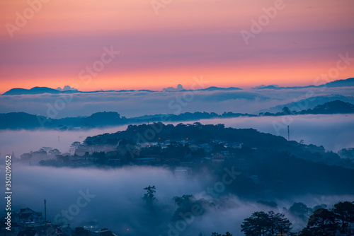 Mountains in fog at beautiful morning in autumn. Landscape with Langbiang mountain valley, low clouds, forest, colorful sky , city illumination at dusk.