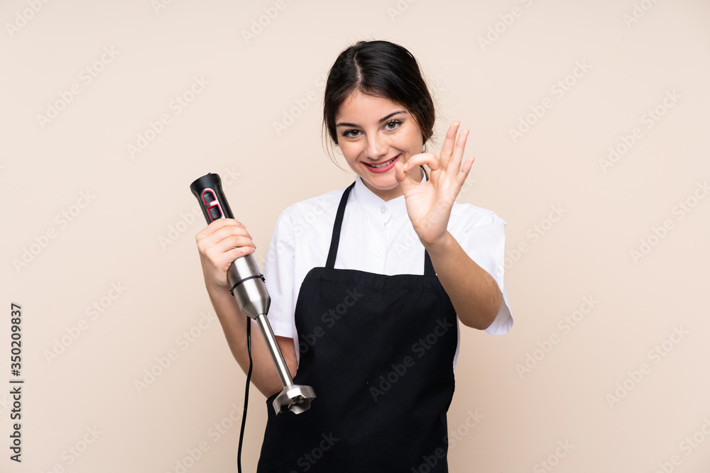 Young woman using hand blender over isolated background showing ok sign with fingers Young woman using hand blender over isolated background showing ok sign with fingers