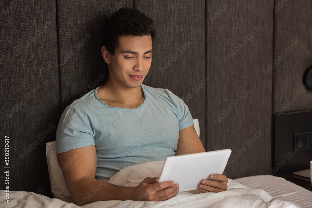 Naklejka premium handsome mixed race man using digital tablet in bed during self isolation