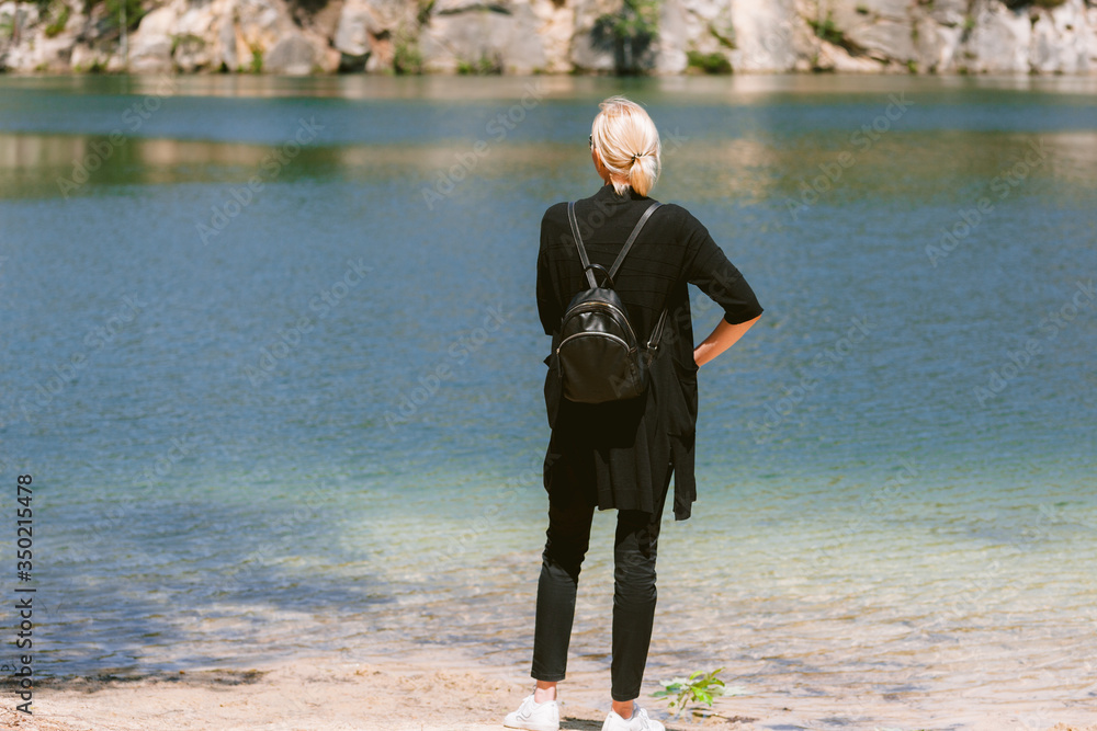 Young pretty blonde woman standing backwards and watching lake in mountainous country landscape. Girl with little backpack outdoors. Backview Young pretty blonde woman standing backwards and watching lake in mountainous country landscape. Girl with little backpack outdoors. Backview