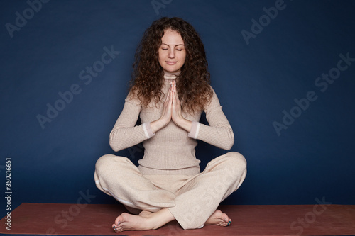 Calm relieved curly haired woman takes deep breath, keeps palms in namaste gesture,  practices yoga or meditation, sitting on mat with crossed legs, bare feet , eyes closed, feeling stress free indoor