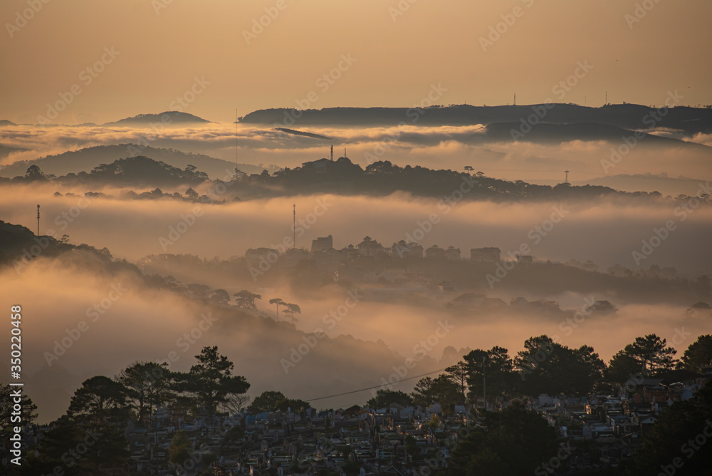 Obraz premium Mountains in fog at beautiful morning in autumn in Dalat city, Vietnam. Landscape with Langbiang mountain valley, low clouds, forest, colorful sky , city illumination at dusk.