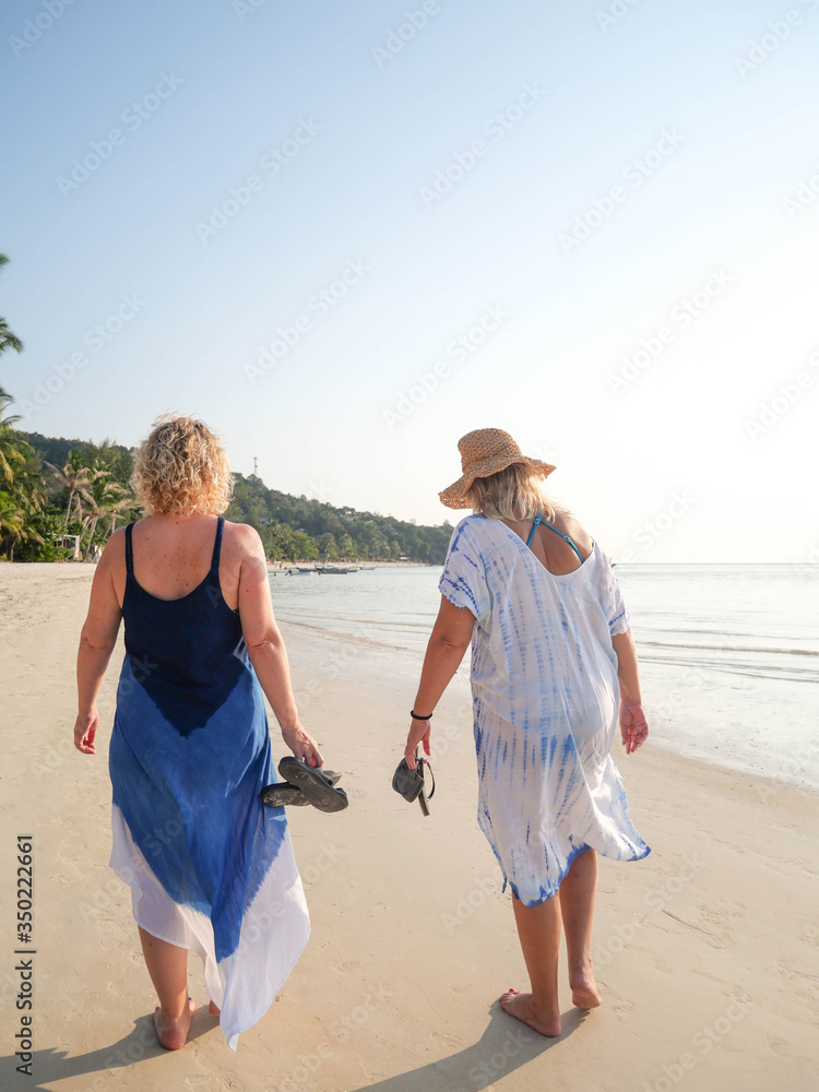 Rear view senior women walking barefoot at sea shore at sunset