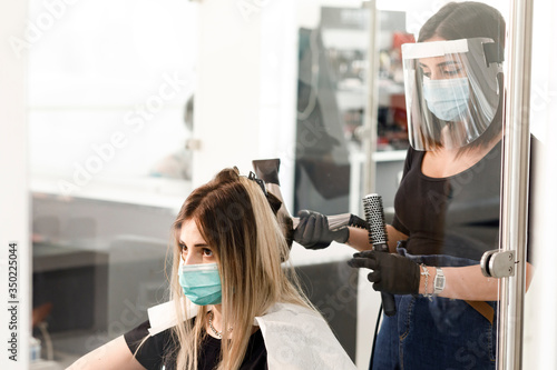 parrucchiera si accinge ad asciugare con un phon i capelli di una di una cliente all'interno di un salone. Entrambi sono equipaggiati di protezioni per evitare contatti e contaminazioni reciproche photo