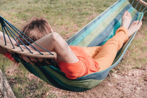 Woman in orange t-shirt and yellow pants relaxing laying in hammock with legs crossed and arms behind head