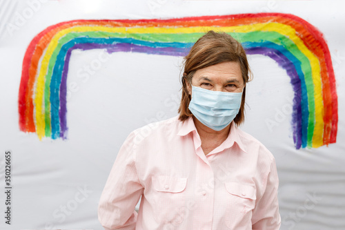 Ritratto di signora anziana che indossa una mascherina chirurgica isolata uno sfondo con un arcobaleno colorato da un bambino appeso su un lenzuolo photo