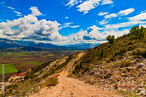 Mountain rocky path in Balkan mountains in Southeren Albania, Saranda, Vlora district in sunny hot summer day. Blue sky, white clouds. View to green farming fields in the valley. photo