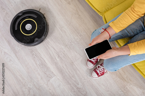 Cropped view of child with smartphone on sofa near robotic vacuum cleaner on floor in living room