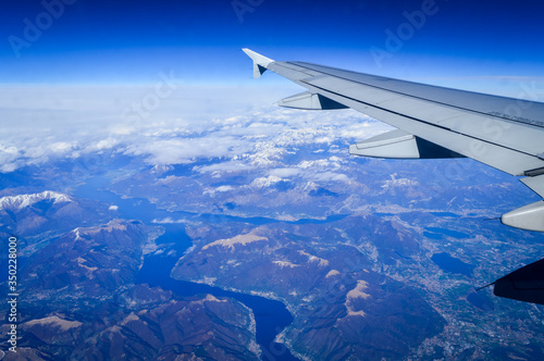 View over the clouds from the porthole of an airplane with plane wing and blue sky over the Como Lake in Italy