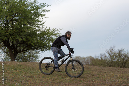Sports brutal bearded guy on a modern mountain bike. Cyclist on the green hills in the spring. © Mountains Hunter