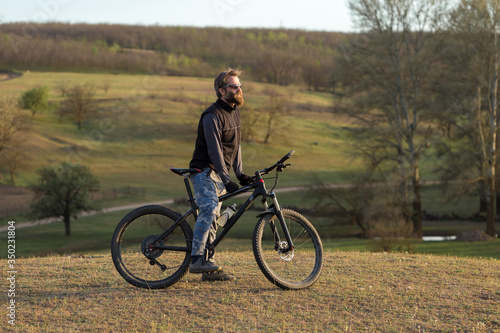 Sports brutal bearded guy on a modern mountain bike. Cyclist on the green hills in the spring.