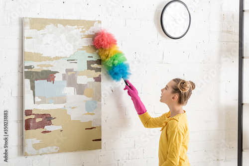 Fotografie Cute child in rubber gloves sweeping away dust from picture with feather duster