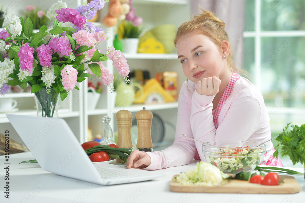 Cute teen girl using laptop while preparing fresh salad on kitchen table Cute teen girl using laptop while preparing fresh salad on kitchen table