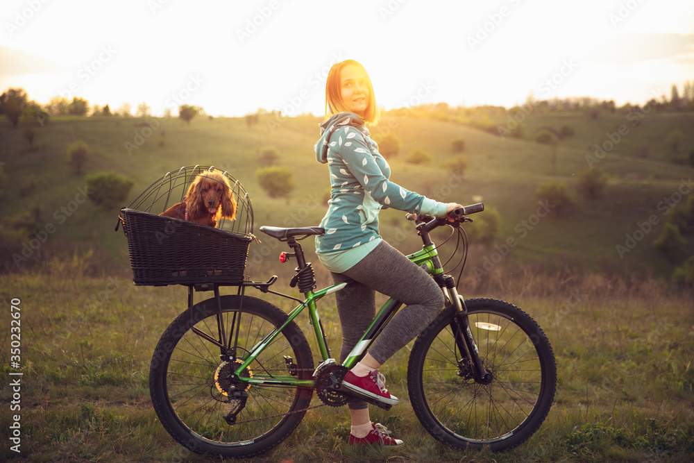 Obraz premium Young woman having fun near countryside park, riding bike, traveling with companion spaniel dog. Calm nature, spring day, positive emotions. Sportive, active leisure activity. Walking together.