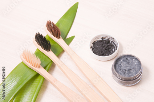 bamboo toothbrushes on a wooden table  focus on a brush with black bristles  homemade charcoal toothpaste in a small glass jar  leaves long plants  eco friendly life style concept.