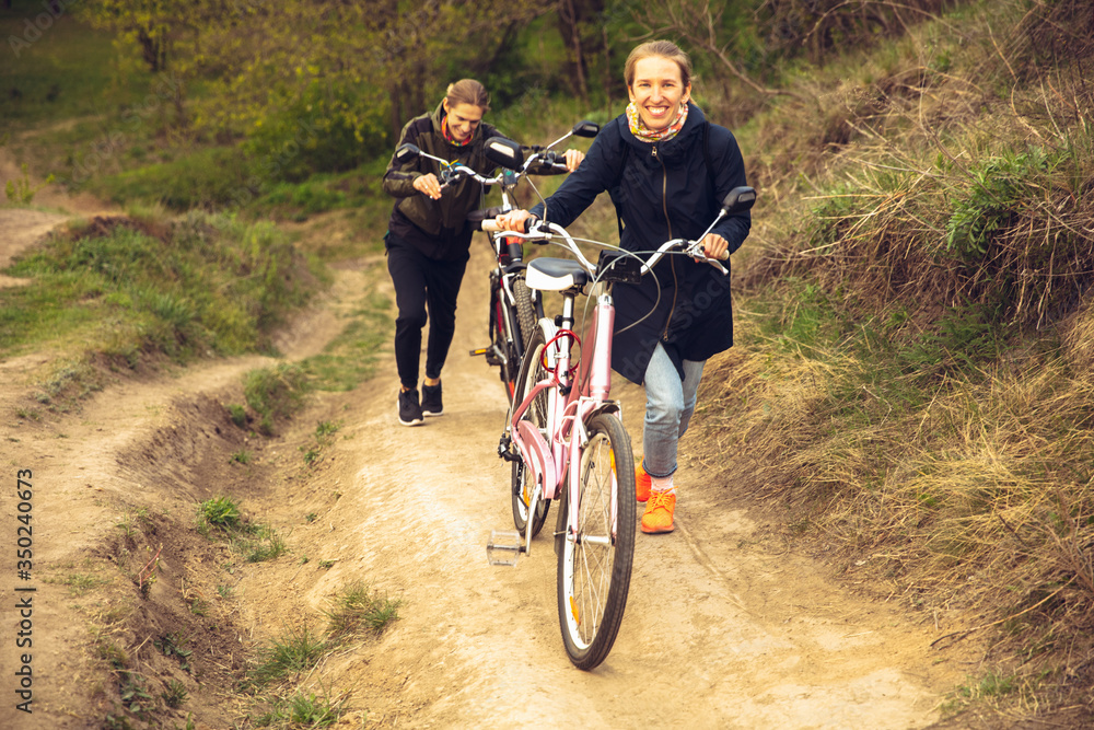 Fototapeta premium Best friends having fun near countryside park, riding bikes, spending time healthy. Calm nature, spring day, positive emotions. Sportive, active leisure activity. Traveling or walking together.