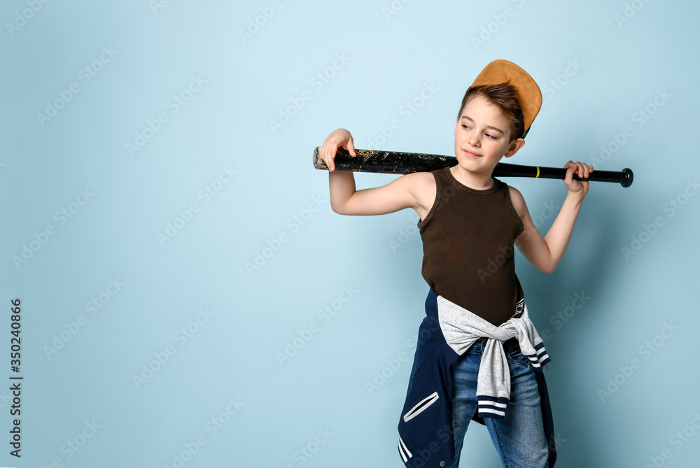 Teenage boy in cap, undershirt, jeans and hoodie tied around his waist. Holding black baseball bat, posing on blue background Teenage boy in cap, undershirt, jeans and hoodie tied around his waist. Holding black baseball bat, posing on blue background