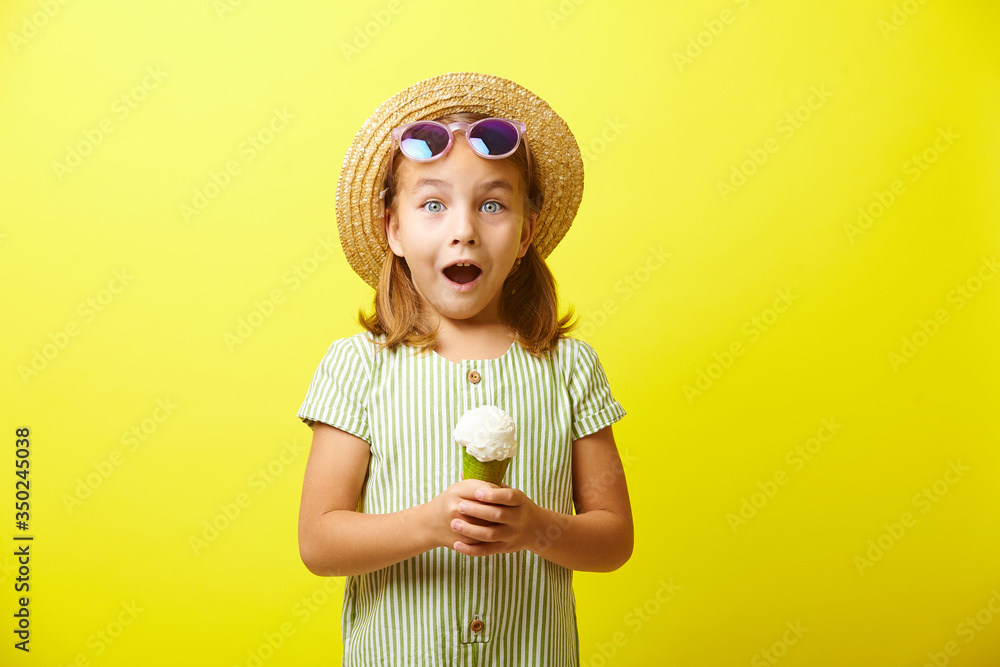 Φωτογραφία Studio portrait of little girl in summer dress, expresses surprise emotion, wears straw hat and sunglasses, holding ice cream on hand, stands over yellow isolated