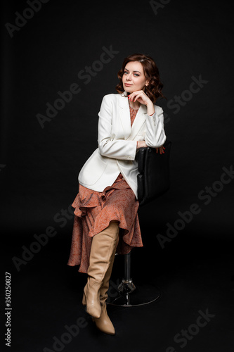 Nice caucasian young female with short dark hair, pretty face in long orange dress, ocher color long boots, white jacket smiles