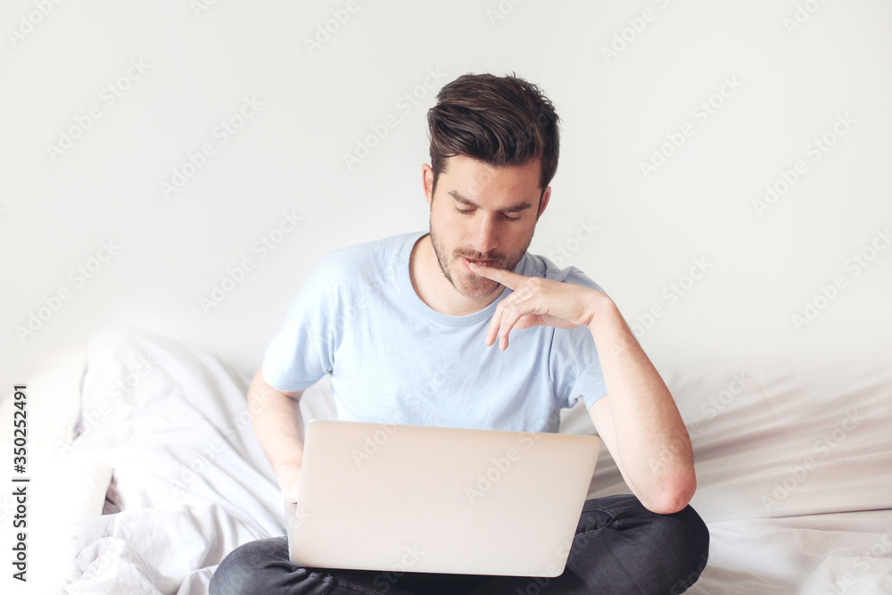 Handsome casual young man in bed with laptop. Solving business troubles in a distance. Concept of work in isolation Handsome casual young man in bed with laptop. Solving business troubles in a distance. Concept of work in isolation
