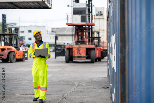 African technician dock worker in protective safety jumpsuit uniform and with hardhat and usel aptop computer at cargo container shipping warehouse. transportation import,export logistic industrial