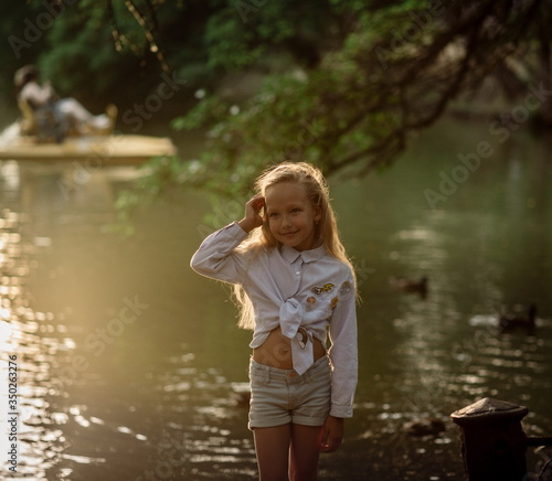Fototapeta Naklejka Na Ścianę i Meble -  A beautiful girl of 7 years old with long blonde hair walks at sunset in a park near a pond on a summer evening