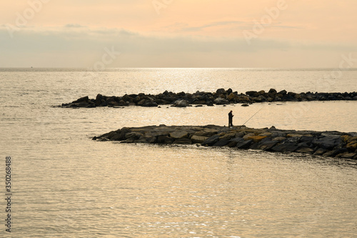 Elevated view of a seascape with the silhouette of a man fishing with a rod on a rocky breakwater at sunset, Italy photo