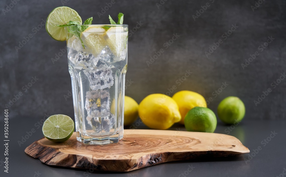 Fototapeta premium Cold refreshing limonade, mojito or gin tonic in glass, with fresh mint and ice cubes, lime and lemon on wooden board and black background. Summer cocktails and drinks concepts. Copy space for text.