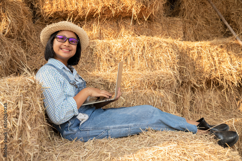 Young girl wear glasses and using a notebook to study agriculture with relax on a haystack in the barn. She has the idea of farming and expanding her father's career as a farmer. Light and shadow. Young girl wear glasses and using a notebook to study agriculture with relax on a haystack in the barn. She has the idea of farming and expanding her father's career as a farmer. Light and shadow.