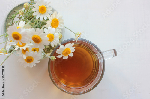 Chamomile herbal tea in glass cup and bouquet of white daisies in a glass vase on the white wooden background with copy space. Top view