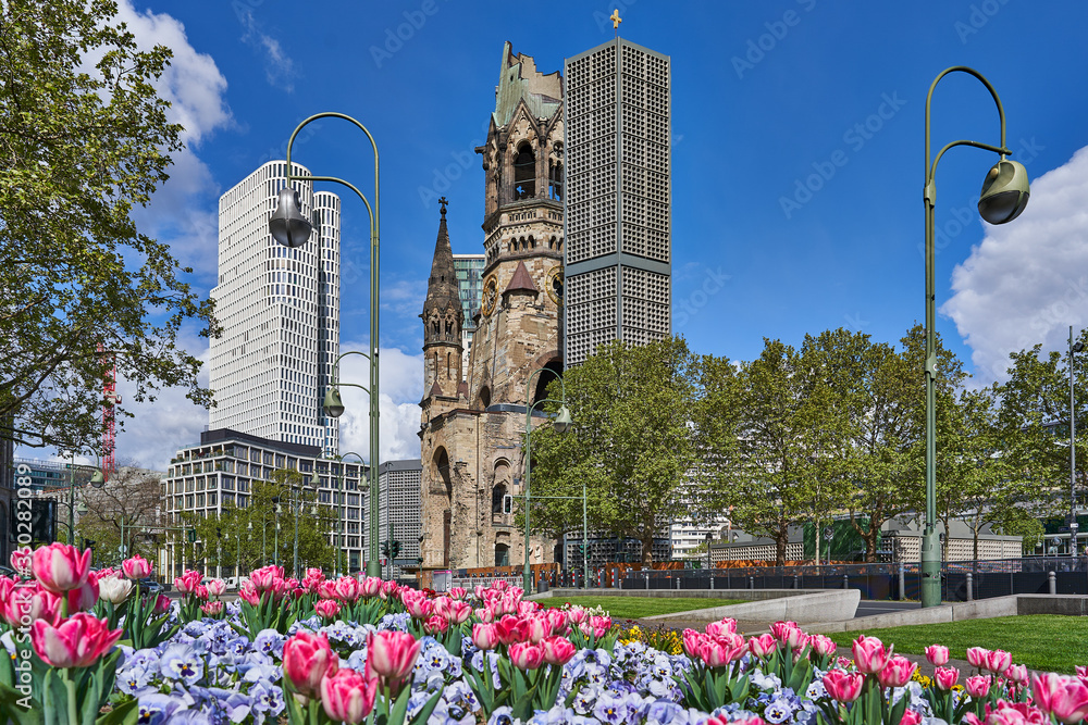 Obraz na płótnie berlin germany, The Kaiser Wilhelm Memorial Church, in GermanKaiser-Wilhelm-Gedächtniskirche, spring time in the city, with a blue sky and colourful flowers, green trees on the road side w salonie