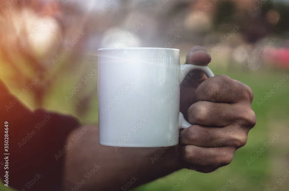 Man holding an empty white cup outdoors, close up. Man's hand holding a porcelain or ceramic mug against nature background. Mockup image, dark vibrant tone, copy space for text. Man holding an empty white cup outdoors, close up. Man's hand holding a porcelain or ceramic mug against nature background. Mockup image, dark vibrant tone, copy space for text.