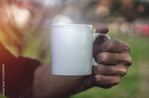 Man holding an empty white cup outdoors, close up. Man's hand holding a porcelain or ceramic mug against nature background. Mockup image, dark vibrant tone, copy space for text.