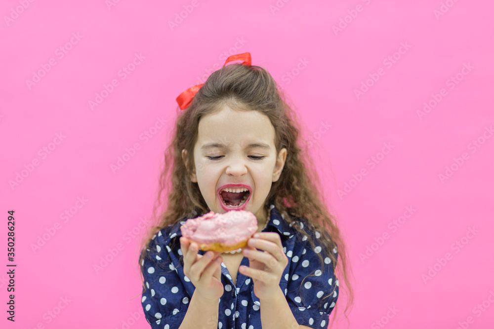 A girl standing over pink background biting a donut.