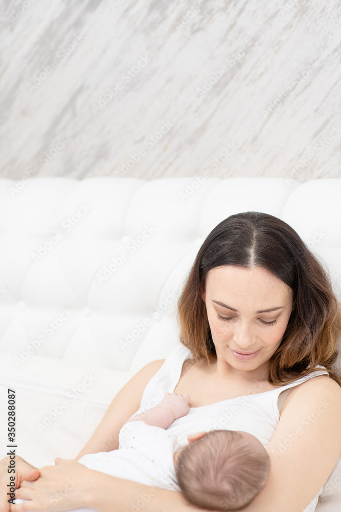 A young mother with a newborn son in her arms looks at the baby with a smile covering her eyes A young mother with a newborn son in her arms looks at the baby with a smile covering her eyes