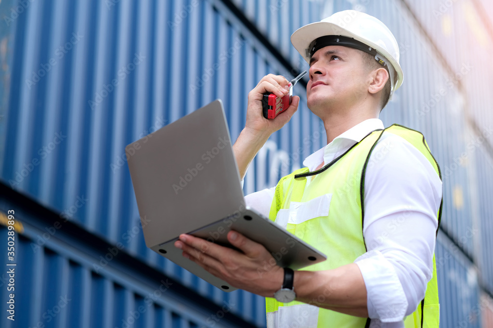 American engineers wear white helmet and holding laptop. Standing at the container shipping area and used radio communication. He talks to another worker. Industrial and shipping business concept.