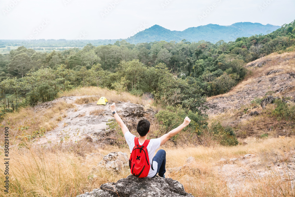 Rear view of young man backpack traveler sit on stone at peak of mountain and had rise arms up to sky. Victory and success concept.