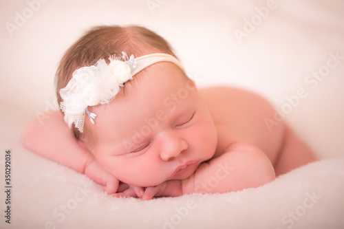 Newborn baby girl, sleeping in a light brown background and lying her head in a tiny pillow. She is wrapped in a tiny blanket and is wearing a flower headband in her head.