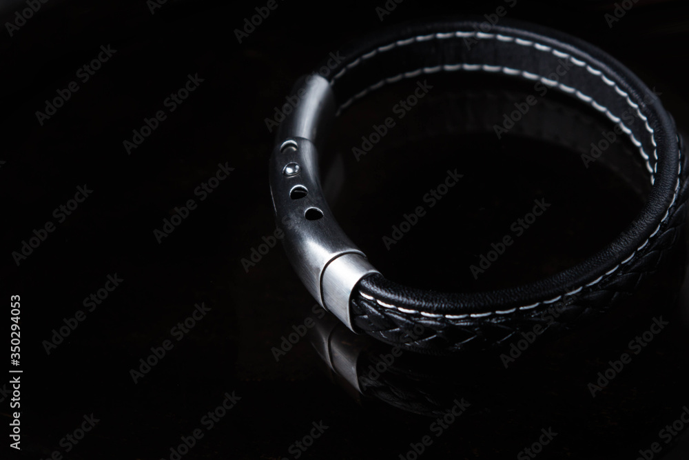 A black leather men's bracelet lies on black glass. It has a metal clasp