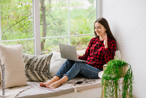 Young beautiful woman sitting on windowsill and using laptop during quarantine. Stay at home. Online communication with friends photo