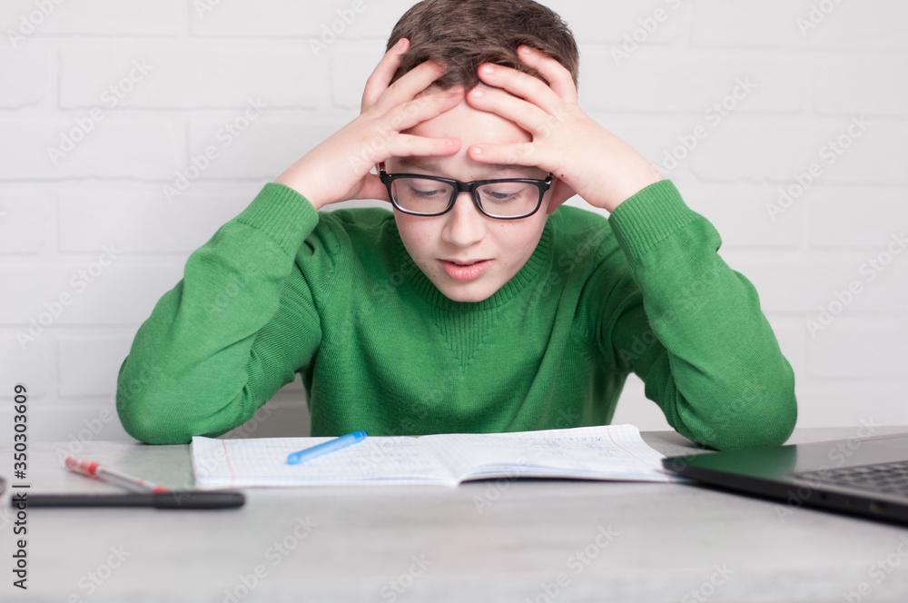 pupil boy is upset of hometasks. touching head both hands sitting at table in light room pupil boy is upset of hometasks. touching head both hands sitting at table in light room