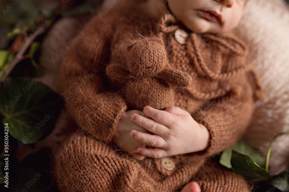 A newborn charming boy, 5 days old, sleeps in a cozy bear outfit and in a  light brown outfit. He sleeps in a basket and holds a tiny teddy bear.  Stock Photo |
